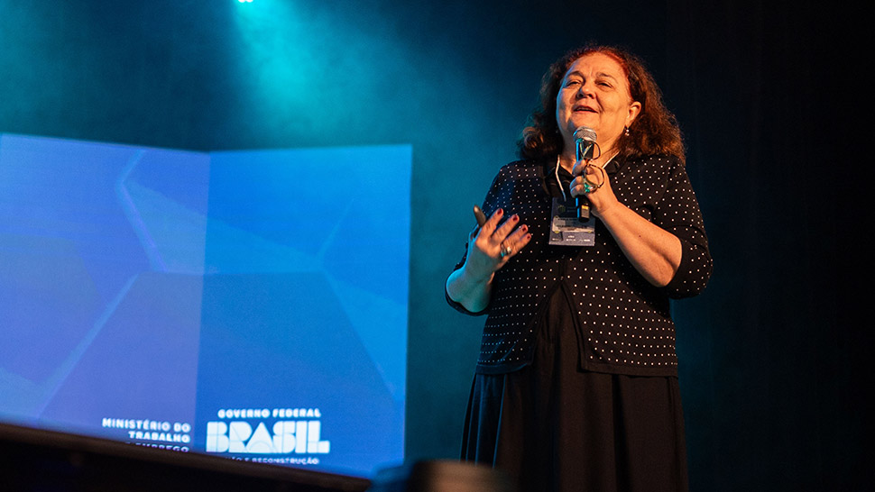 Paula Montagner, mulher de pele clara, cabelos castanhos escuros e ondulados, veste blusa preta de bolinhas brancas e saia preta. Ela está em pé no palco do Teatro CIEE, segurando um microfone e apresentando dados em uma grande tela de LED ao fundo. A projeção exibe tópicos como estágio, aprendizagem, automação, inteligência artificial e políticas de educação para jovens entre 14 e 24 anos. À esquerda e à direita da tela, estão os logos do Ministério do Trabalho e Emprego e do Governo Federal. A iluminação do palco é esverdeada.
