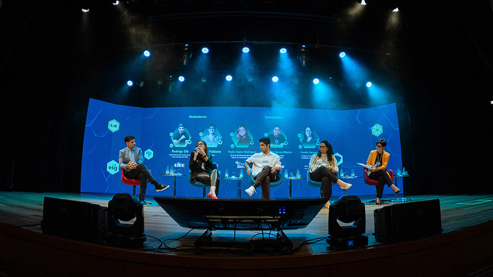 Cinco pessoas estão sentadas em poltronas coloridas no palco do Teatro CIEE, participando da mesa de conversa do evento Empregabilidade Jovem Brasil 2025. À esquerda, Rodrigo Dib (CIEE) e Paula Fabiani (IDIS) atuam como mediadores. À direita, os convidados: Keyla Copes Rodrigues (Fundação Sicredi), Wallace da Neves Ribeiro (Ambev) e Camila Valverde (Fundação ArcelorMittal) participam da discussão. No telão ao fundo, aparecem os nomes e fotos dos participantes, acompanhados das logomarcas das instituições representadas. A plateia acompanha atentamente. O palco tem iluminação em tons azulados e verdes, e há garrafas de água e papéis sobre as mesas laterais.