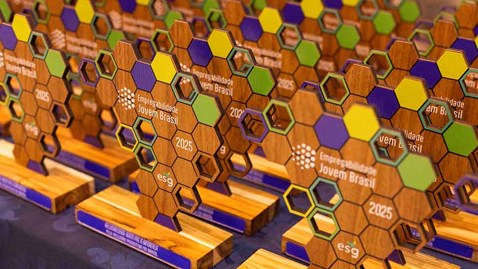 imagem em close de um dos troféus do Empregabilidade Jovem Brasil 2025, apoiado sobre uma toalha azul escura com arabescos. O troféu tem formato inspirado no mapa do Brasil, composto por hexágonos de madeira com recortes coloridos em roxo, verde e amarelo. Estão impressos os dizeres “Empregabilidade Jovem Brasil”, o ano “2025” e a sigla “esg”. A base é de madeira clara com plaqueta roxa, destacando o nome da empresa premiada. Ao fundo, desfocados, aparecem outros troféus e uma pessoa com óculos.