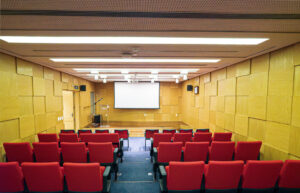 Foto do auditório com paredes amarelas e cadeiras vermelhas. Há um telão no fundo
