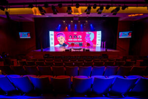 Foto do Teatro CIEE visto de cima. São diversas cadeiras voltadas para um grande palco com um grande telão