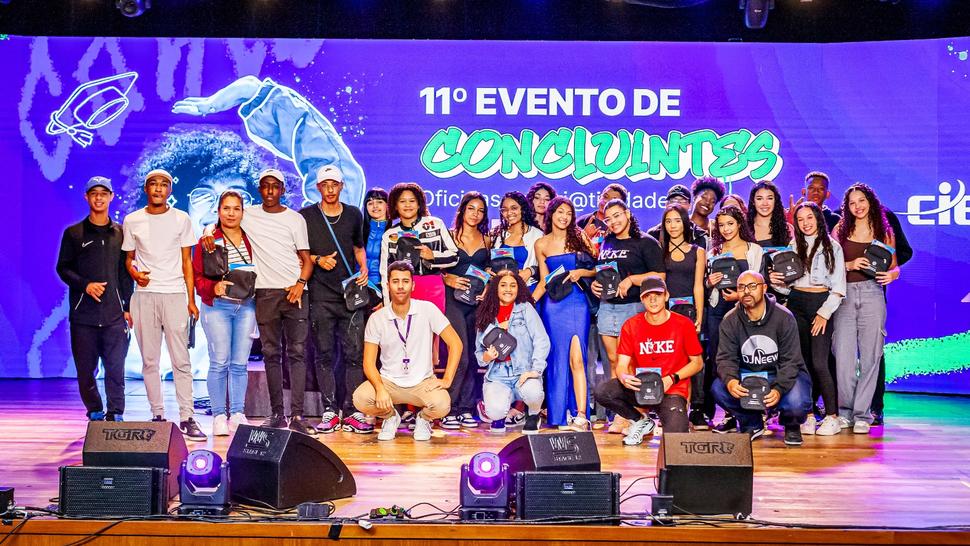 Jovens em um palco, posando para foto, atrás há um telão com o nome do evento e fundo roxo