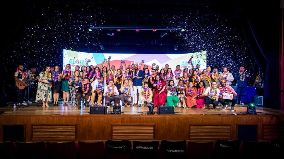 Diversas pessoas com prêmios em mão, em cima de um palco, com confetes
