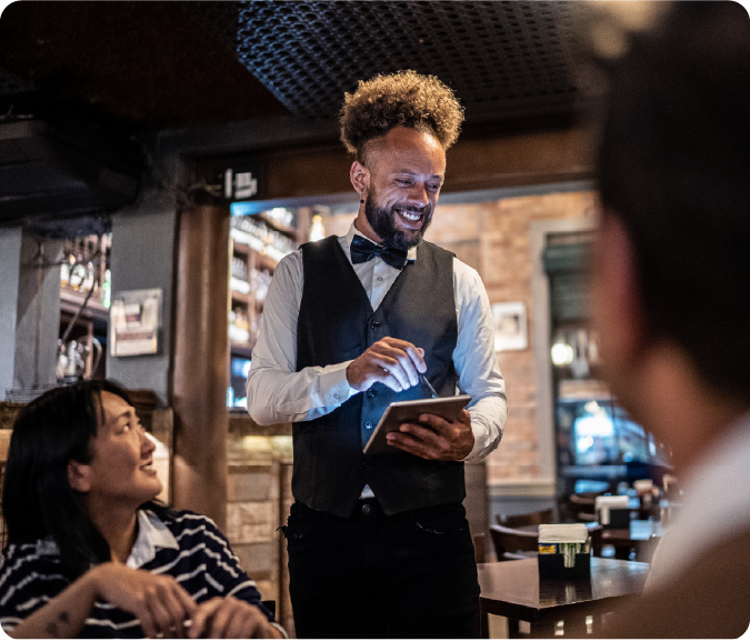 Foto de um garçom negro do Outback anotando o pedido de um casal sentado a mesa