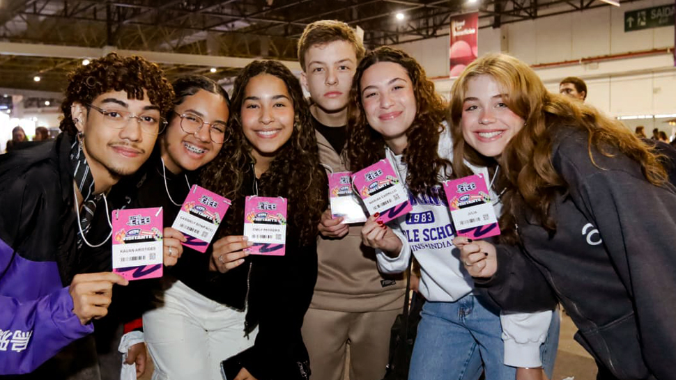 Seis jovens posam sorrindo para a câmera dentro do pavilhão da EXPO CIEE 2025. Eles seguram seus crachás de identificação do evento e parecem animados. Ao fundo, há banners pendurados no teto e outras pessoas circulando pelo espaço.