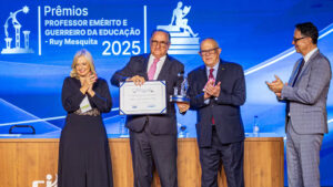 Quatro pessoas em cima do palco, sendo três homens e uma mulher. Todos usam vestes sociais e o telão ao fundo é azul. Antonio Palma segura seu prêmio de forma orgulhosa levantando ao público.