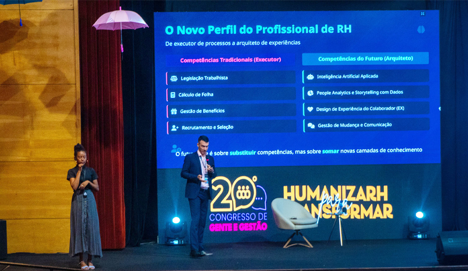 Auditório cheio com pessoas sentadas assistindo a uma palestra no palco. À direita, um homem de terno azul apresenta com microfone em mãos diante de um telão que exibe o título “O Novo Perfil do Profissional de RH” e uma tabela comparando competências tradicionais e do futuro. À esquerda, uma intérprete de Libras acompanha a apresentação.
