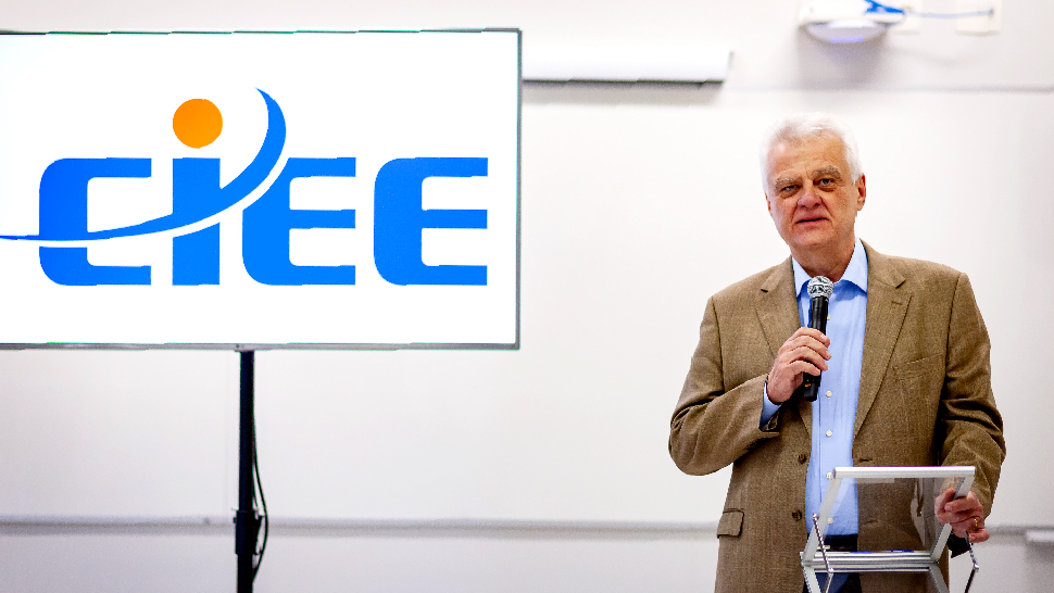 Homem de cabelo branco, vestindo blazer ferrugem com blusa social azul. Ele fala ao microfone em um púlpito. Ao seu lado uma TV mostra o logo do CIEE.