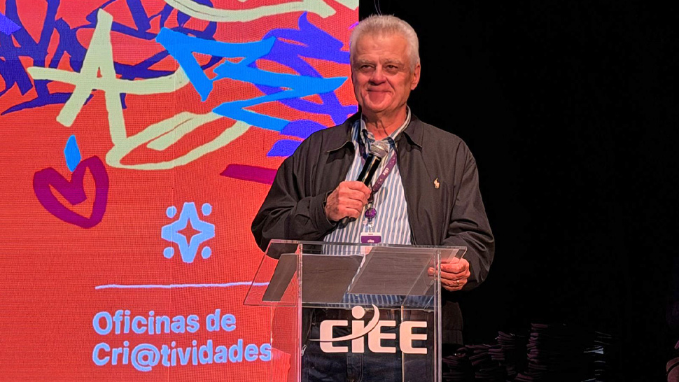 Homem branco de cabelos grisalhos, vestindo jaqueta preta e segurando um microfone em um púlpito transparente com o logo do CIEE. Ele está em um palco, discursando diante de um painel colorido com o texto "Oficinas de Cri@tividades".