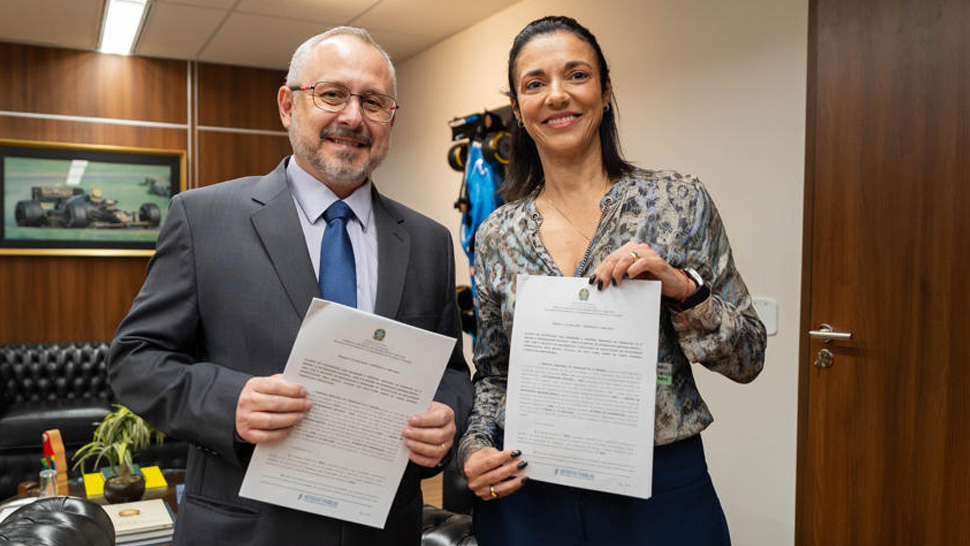 Duas pessoas em ambiente institucional posam lado a lado, sorrindo e segurando documentos oficiais assinados. À esquerda, Marcelo Gallo, Superintendente Executivo do CIEE, e à direita, uma representante da instituição. A cena ocorre em um escritório com móveis de madeira, sofá ao fundo e decoração formal, indicando a formalização de um acordo.