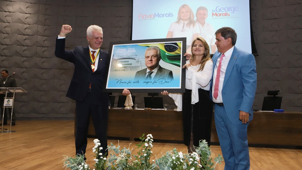Fotografia colorida em plano inteiro tirada em um auditório. À esquerda, Humberto Casagrande, um homem de cabelos brancos vestindo terno escuro e medalha no pescoço, ergue o braço direito com o punho cerrado e sorri. No centro e à direita, a deputada Flávia Morais, vestindo blusa branca e saia escura, e o deputado Dr. George Morais, de terno azul claro, seguram juntos um grande quadro emoldurado.