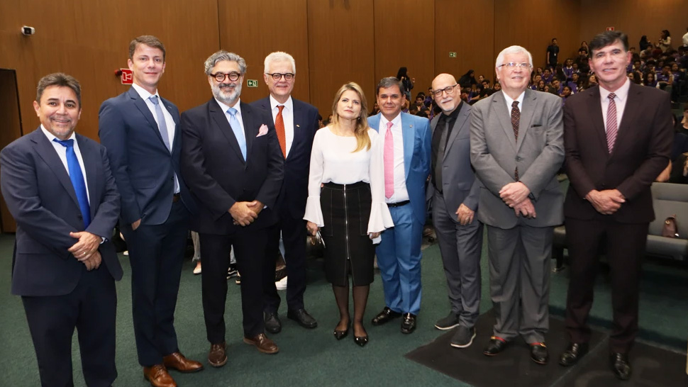 Grupo de oito pessoas posando para foto em um auditório. Todos estão vestidos com roupas sociais, em sua maioria ternos escuros e gravatas. No centro da imagem há uma mulher com blusa branca e saia preta. Ao fundo, aparecem poltronas ocupadas por público e paredes de madeira iluminadas por luz interna.