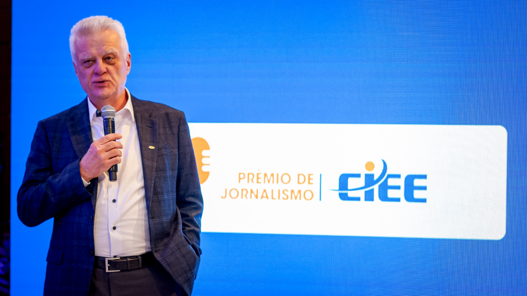 Humberto Casagrande, CEO do CIEE, está em pé segurando um microfone enquanto faz um discurso durante o lançamento do Prêmio CIEE de Jornalismo. Ele veste blazer azul escuro e camisa branca. Ao fundo, um painel de LED azul exibe o logotipo do Prêmio CIEE de Jornalismo e do CIEE.
