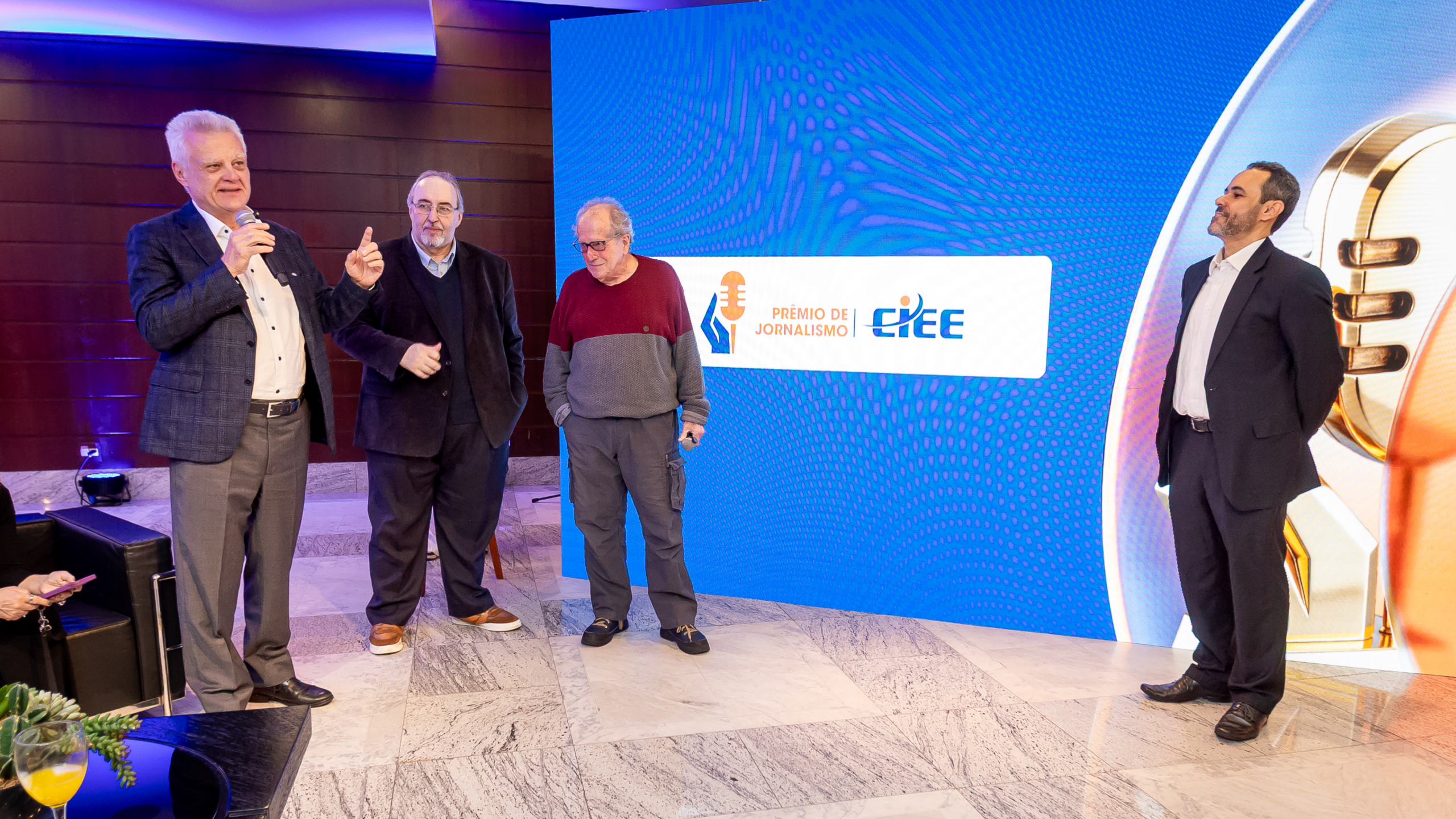 Quatro homens estão em pé durante o lançamento do Prêmio CIEE de Jornalismo, em um ambiente de evento. À esquerda, um deles fala ao microfone enquanto gesticula. Ao fundo, um grande painel de LED azul exibe a identidade visual do Prêmio CIEE de Jornalismo e o logotipo do CIEE. À direita, outro participante observa a apresentação. O espaço possui piso de mármore e iluminação interna.