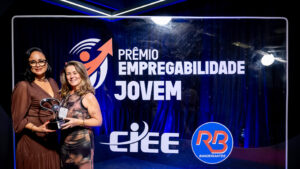Foto de um evento de premiação formal em um estúdio ou palco. O fundo é um painel de evento retroiluminado e texturizado, de cor azul royal profundo, com logotipos e texto claros.