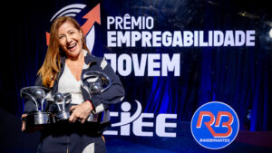 Foto de um evento de premiação formal em um estúdio ou palco. O fundo é um painel de evento retroiluminado e texturizado, de cor azul royal profundo, com logotipos e texto claros.