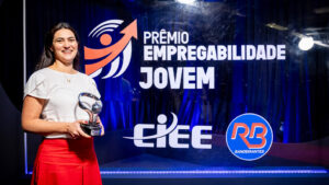 Foto de um evento de premiação formal em um estúdio ou palco. O fundo é um painel de evento retroiluminado e texturizado, de cor azul royal profundo, com logotipos e texto claros.