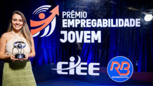 Foto de um evento de premiação formal em um estúdio ou palco. O fundo é um painel de evento retroiluminado e texturizado, de cor azul royal profundo, com logotipos e texto claros.