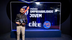 Foto de um evento de premiação formal em um estúdio ou palco. O fundo é um painel de evento retroiluminado e texturizado, de cor azul royal profundo, com logotipos e texto claros.