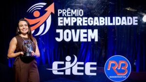 Foto de um evento de premiação formal em um estúdio ou palco. O fundo é um painel de evento retroiluminado e texturizado, de cor azul royal profundo, com logotipos e texto claros.