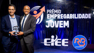 Foto de um evento de premiação formal em um estúdio ou palco. O fundo é um painel de evento retroiluminado e texturizado, de cor azul royal profundo, com logotipos e texto claros.