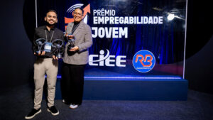 Foto de um evento de premiação formal em um estúdio ou palco. O fundo é um painel de evento retroiluminado e texturizado, de cor azul royal profundo, com logotipos e texto claros.
