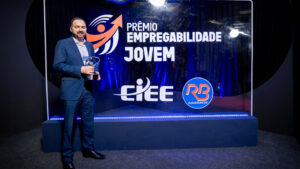 Foto de um evento de premiação formal em um estúdio ou palco. O fundo é um painel de evento retroiluminado e texturizado, de cor azul royal profundo, com logotipos e texto claros.