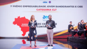 O fundo é um grande painel de LED curvo e texturizado em tons de cinza claro com acentos sutis de vermelho e amarelo no canto superior esquerdo. No canto superior direito, há o logotipo e o texto do evento em destaque: um ícone estilizado de seta vermelha e azul e o texto "PRÊMIO EMPREGABILIDADE JOVEM" em letras grandes, pretas e vermelhas em negrito. Logo abaixo, no lado direito, estão os logotipos institucionais adicionais: "CIEE" (em azul) e o logotipo circular "RB Bandeirantes" (em azul e vermelho). O chão do palco é uma superfície de acrílico preto reflexivo, espelhando sutilmente a imagem. Esta estrutura de palco e painel de fundo são os mesmos para todas as imagens.
