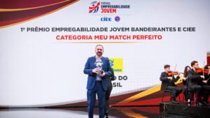O fundo é um grande painel de LED curvo e texturizado em tons de cinza claro com acentos sutis de vermelho e amarelo no canto superior esquerdo. No canto superior direito, há o logotipo e o texto do evento em destaque: um ícone estilizado de seta vermelha e azul e o texto "PRÊMIO EMPREGABILIDADE JOVEM" em letras grandes, pretas e vermelhas em negrito. Logo abaixo, no lado direito, estão os logotipos institucionais adicionais: "CIEE" (em azul) e o logotipo circular "RB Bandeirantes" (em azul e vermelho). O chão do palco é uma superfície de acrílico preto reflexivo, espelhando sutilmente a imagem. Esta estrutura de palco e painel de fundo são os mesmos para todas as imagens.