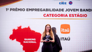 O fundo é um grande painel de LED curvo e texturizado em tons de cinza claro com acentos sutis de vermelho e amarelo no canto superior esquerdo. No canto superior direito, há o logotipo e o texto do evento em destaque: um ícone estilizado de seta vermelha e azul e o texto "PRÊMIO EMPREGABILIDADE JOVEM" em letras grandes, pretas e vermelhas em negrito. Logo abaixo, no lado direito, estão os logotipos institucionais adicionais: "CIEE" (em azul) e o logotipo circular "RB Bandeirantes" (em azul e vermelho). O chão do palco é uma superfície de acrílico preto reflexivo, espelhando sutilmente a imagem. Esta estrutura de palco e painel de fundo são os mesmos para todas as imagens.