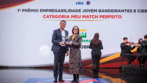 O fundo é um grande painel de LED curvo e texturizado em tons de cinza claro com acentos sutis de vermelho e amarelo no canto superior esquerdo. No canto superior direito, há o logotipo e o texto do evento em destaque: um ícone estilizado de seta vermelha e azul e o texto "PRÊMIO EMPREGABILIDADE JOVEM" em letras grandes, pretas e vermelhas em negrito. Logo abaixo, no lado direito, estão os logotipos institucionais adicionais: "CIEE" (em azul) e o logotipo circular "RB Bandeirantes" (em azul e vermelho). O chão do palco é uma superfície de acrílico preto reflexivo, espelhando sutilmente a imagem. Esta estrutura de palco e painel de fundo são os mesmos para todas as imagens.