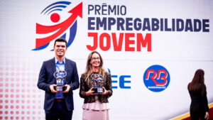 O fundo é um grande painel de LED curvo e texturizado em tons de cinza claro com acentos sutis de vermelho e amarelo no canto superior esquerdo. No canto superior direito, há o logotipo e o texto do evento em destaque: um ícone estilizado de seta vermelha e azul e o texto "PRÊMIO EMPREGABILIDADE JOVEM" em letras grandes, pretas e vermelhas em negrito. Logo abaixo, no lado direito, estão os logotipos institucionais adicionais: "CIEE" (em azul) e o logotipo circular "RB Bandeirantes" (em azul e vermelho). O chão do palco é uma superfície de acrílico preto reflexivo, espelhando sutilmente a imagem. Esta estrutura de palco e painel de fundo são os mesmos para todas as imagens.