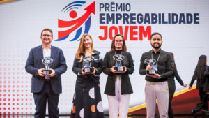 O fundo é um grande painel de LED curvo e texturizado em tons de cinza claro com acentos sutis de vermelho e amarelo no canto superior esquerdo. No canto superior direito, há o logotipo e o texto do evento em destaque: um ícone estilizado de seta vermelha e azul e o texto "PRÊMIO EMPREGABILIDADE JOVEM" em letras grandes, pretas e vermelhas em negrito. Logo abaixo, no lado direito, estão os logotipos institucionais adicionais: "CIEE" (em azul) e o logotipo circular "RB Bandeirantes" (em azul e vermelho). O chão do palco é uma superfície de acrílico preto reflexivo, espelhando sutilmente a imagem. Esta estrutura de palco e painel de fundo são os mesmos para todas as imagens.