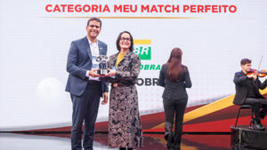 O fundo é um grande painel de LED curvo e texturizado em tons de cinza claro com acentos sutis de vermelho e amarelo no canto superior esquerdo. No canto superior direito, há o logotipo e o texto do evento em destaque: um ícone estilizado de seta vermelha e azul e o texto "PRÊMIO EMPREGABILIDADE JOVEM" em letras grandes, pretas e vermelhas em negrito. Logo abaixo, no lado direito, estão os logotipos institucionais adicionais: "CIEE" (em azul) e o logotipo circular "RB Bandeirantes" (em azul e vermelho). O chão do palco é uma superfície de acrílico preto reflexivo, espelhando sutilmente a imagem. Esta estrutura de palco e painel de fundo são os mesmos para todas as imagens.