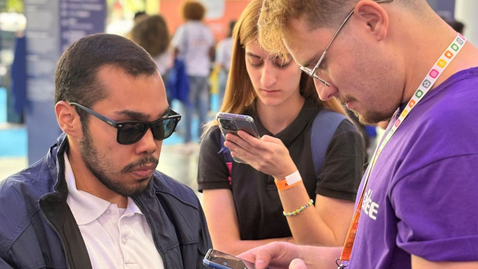 Três jovens estão em um ambiente de evento movimentado, como uma conferência ou feira. No primeiro plano, à direita, um homem loiro de óculos e camisa roxa, usando um crachá de evento, segura e olha para a tela de um smartphone. À esquerda, um homem de cabelo raspado, barba rala e óculos de sol escuros, vestindo uma jaqueta azul sobre uma camisa branca, está concentrado e olha para a mesma tela. Ao fundo, entre os dois, uma jovem loira de camisa preta está focada no seu próprio smartphone. O fundo está desfocado, mostrando outras pessoas e estruturas de estandes de evento.