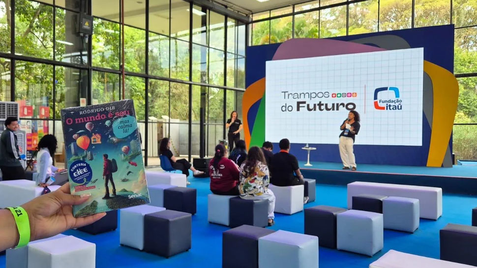 Esta imagem mostra uma mão segurando um livro em primeiro plano, com um palco de conferência ao fundo. A mão, visível no lado esquerdo, segura o livro intitulado "O mundo é seu", de Rodrigo Dib. A capa do livro apresenta ilustrações coloridas de balões, nuvens e um personagem em pé, junto com selos da 'CIEE' e 'Senac'. O plano de fundo é um espaço de conferência moderno e bem iluminado, com piso azul vibrante e grandes janelas de vidro do chão ao teto, revelando uma vista para o jardim. No palco, uma grande tela de LED exibe o texto 'Trampos do Futuro', os logotipos da Fundação Itaú e CIEE, e outras formas coloridas. Duas pessoas estão no palco: uma mulher de cabelo escuro e camisa preta está apresentando no lado esquerdo, enquanto um homem de cabelo curto está em pé no centro, gesticulando. Uma plateia está sentada em bancos de cubo cinza e azul, olhando para o palco. Há também outros participantes em pé ao longe. O ambiente é dinâmico e focado na apresentação.